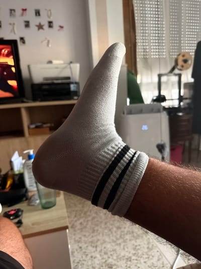 The image shows a close-up of a person's right foot wearing a grey sock with black stripes. The foot is extended upwards and to the left, with the heel facing the viewer. The sock appears to be made of a ribbed material, and the black stripes are positioned around the ankle area. The person's leg, covered in dark hair, is visible below the sock.
In the background, a wooden shelf unit is partially visible, holding a printer and what appears to be a television screen showing a brightly colored image. To the right, a white portable air conditioner unit is present, with a blue digital display showing a snowflake icon. Behind the air conditioner, sheer curtains with a circular pattern cover a window, and a wooden chair with a red cushion is visible. The environment suggests an indoor residential setting.
The image is focused on the foot and leg, with the background elements out of focus. The lighting appears to be artificial, casting shadows and highlighting the texture of the sock and the hair on the leg. There is no information in the image to indicate the location as Castillo Bajo-conejito, Spain.