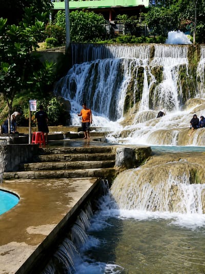 The image depicts a multi-tiered waterfall with several people present in the vicinity. In the foreground, a man in an orange t-shirt and black shorts stands on a wet concrete platform next to a set of stone steps. To his left, another man in a black shirt and shorts stands by a red stool, near a sign with text. Further left, two figures are visible near a blue pool of water.
The waterfall itself cascades over rocky, moss-covered formations, with white water tumbling down into pools of clear, blue-green water. In the middle ground, several people are seated on the rocks near the water. Two women wearing hijabs are visible on the right, and another person is partially obscured behind them.
The background is filled with lush green trees and vegetation, and a building with a green roof is visible at the top. The sun is shining, casting shadows and highlighting the water and rocks. The overall scene suggests a natural park or tourist attraction with recreational water features. This location is identified as Silaiyajae, Indonesia.