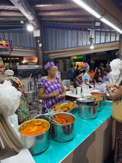 A woman in a purple and white dress stands behind a food stall with various pots of food. The pots contain yellow and orange colored food. Another woman is visible in the lower part of the image. A line of people stands beyond the stall. A Subway restaurant is visible in the background. The area is covered with wooden beams and blue painted metal structures.