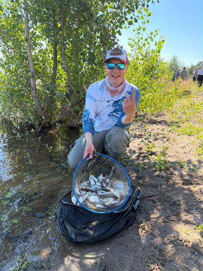 A Caucasian man is kneeling on a sandy bank next to a body of water. He is wearing sunglasses, a patterned long-sleeved shirt, grey pants, and a baseball cap. He is holding a fishing net filled with fish and has his fingers held up in a peace sign. The water is murky and there are trees and vegetation along the bank. Other people can be seen in the background, some walking and others standing. The sun is shining brightly, casting shadows on the ground. The location is Kambarka, Russia.