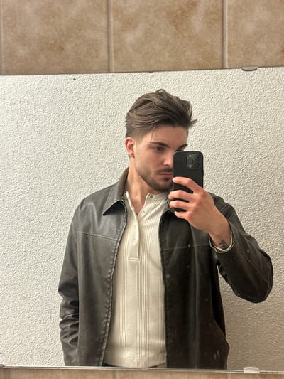 The scene is a mirror reflection of a male individual taking a selfie indoors in Volketswil, Switzerland. The subject is a male with brown hair styled back and a trimmed beard. He is wearing a dark brown, zippered jacket, unzipped, over a light beige, ribbed Henley shirt with a three-button placket. A dark-colored tattoo is visible on his left wrist. He holds a black smartphone, featuring a multi-lens camera array, in his right hand, positioned to photograph his reflection. The visible background consists of a textured, off-white wall in the lower section and a horizontal band of light brown, mottled ceramic tiles above it.
