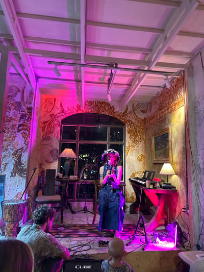 The image depicts an indoor performance space illuminated by pink and purple lighting. A woman in a blue dress and a floral hat is singing into a microphone on a stage. To her right is a keyboard and a small table with a lamp and decorations. In the foreground, a man with curly hair is facing away from the camera, and a child is looking towards the stage. The room features walls with elaborate murals and a large window overlooking an outdoor area. The ceiling has exposed beams. A djembe drum is visible on the left side of the stage. The environment suggests a bohemian or artistic venue. The presence of the child and adult in the foreground indicates an audience. The location is identified as Ko Pha-ngan District, Thailand.