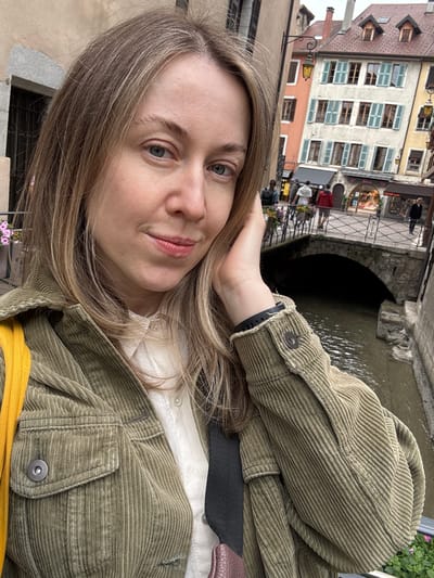 A young woman with blonde hair and blue eyes takes a selfie in Annecy, France. She is wearing a green corduroy jacket over a white shirt. In the background, there is a canal with a stone bridge and colorful buildings typical of the city's old town. People are visible crossing the bridge and walking along the canal. The weather appears overcast.