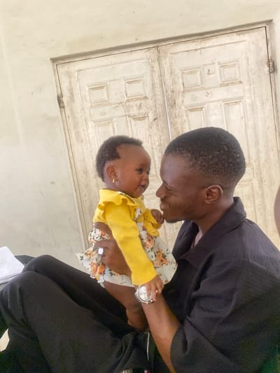 In this image, a Black man is holding a baby. The man is wearing a black shirt and black pants. He is leaning down and smiling at the baby. The baby is wearing a yellow long-sleeved shirt and a floral-patterned dress. The baby is looking up at the man and appears to be smiling. The background of the image consists of a white wooden door with panels. The lighting suggests it could be daytime. The overall scene depicts a tender moment between a father and his child. There is no visible text in the image.