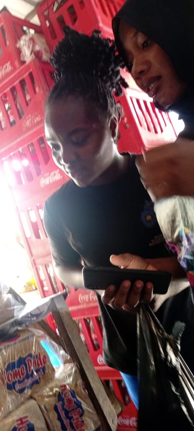 In a dimly lit indoor setting, likely a market stall in Kwachiri, Nigeria, two individuals are captured in a close-up shot. The foreground is dominated by a stack of red Coca-Cola crates, some of which are partially obscured by a wooden shelf holding packages of "Home Pride" bread.
The primary subject is a woman in the foreground, with her head tilted downwards, focusing on a black smartphone she holds in her right hand. She has braided hair styled in a bun and wears a black t-shirt with a faint floral design. To her right, another woman, partially visible and wearing a black headscarf, looks towards the camera. Her gaze is directed upwards, and her mouth is slightly open, suggesting she might be speaking.
The background is filled with more red Coca-Cola crates stacked high, creating a sense of depth and a busy commercial environment. The lighting is uneven, with a strong light source coming from the left, casting highlights on the subjects and the crates. The overall impression is a candid moment captured amidst the daily commerce of a Nigerian market.