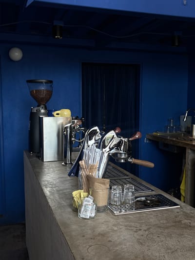 The image depicts a coffee counter in San Pedro Garza García, Mexico. The counter is made of concrete and is set against a vibrant blue wall. On the counter, there is a coffee grinder filled with beans, a small yellow mug, a chrome espresso machine, and various coffee-making accessories such as straws, wooden stirrers, sugar packets, and two empty glasses. To the right of the counter, a sink area is visible with stacked glasses and a faucet. The overall scene suggests a functional and well-equipped coffee preparation area.