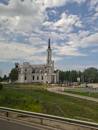 The image shows a white building with a tall spire, resembling a church or a public building, situated in Dybów Kolonia, Poland. The building has a distinctive architectural style with multiple windows and decorative elements. It is surrounded by green grass and trees, with a road and a fence visible to the right. The sky is filled with white clouds against a blue backdrop, suggesting it's daytime with partly cloudy weather. There are also power lines visible in the distance, indicating a developed area. The image appears to be taken from a moving vehicle, as suggested by the angle and the blur in the foreground. No people are clearly visible, and the overall impression is one of a serene landscape with a prominent architectural structure. There is no visible text in the image.