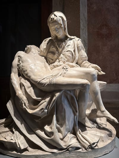 The image displays a large, vertical, white marble sculpture featuring multiple figures, characteristic of a Pieta scene. A central female figure, likely Mary, with a veiled head and a sorrowful expression, supports the limp, deceased body of a male figure, presumably Jesus Christ. His bare torso and splayed limbs are visible, emphasizing his lifeless state. The female figure's hands are delicately placed, holding him in a posture of profound grief. Additional figures are partially visible in the composition, including a hand reaching up near Christ's leg and a larger figure behind Mary.
The sculpture exhibits intricate carving, particularly in the flowing and deeply folded drapery that envelops the figures and forms the base. Anatomical details are finely rendered on the male figure, contrasting with the soft, elegant lines of the female's attire.
The sculpture is illuminated by strong, directional light from the upper left, creating dramatic contrasts of light and shadow that emphasize its three-dimensional quality and texture. The polished marble surfaces reflect this light, giving the sculpture a luminous appearance.
The background consists of dark reddish-brown marble walls with a horizontal band of lighter, veined marble, suggesting an interior setting within a grand building. The lower-left background is very dark, possibly a recessed alcove.
Faint, partially obscured text is visible on a band around the female figure's head covering. While largely illegible, fragmented words such as "SACRUM" and "AGNOSTI STUD..." can be discerned. The overall scene conveys intense emotion, typical of religious art found in places like Città del Vaticano, Vatican City.