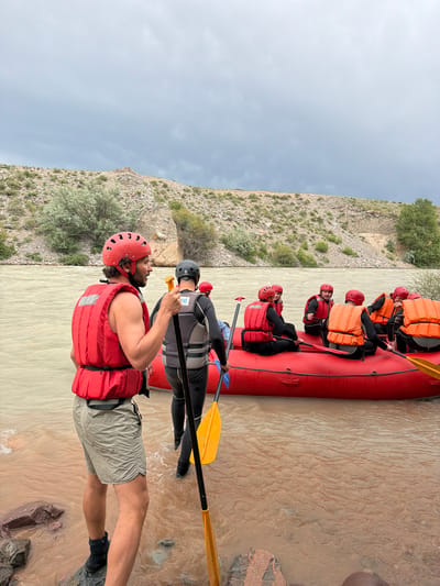 This media file shows a group of people in a red raft on a muddy river, possibly Sasyk-Bulak, Kyrgyzstan. The people are wearing life vests and helmets, indicating they are participating in a water sport like whitewater rafting. The raft is filled with individuals, and some are holding paddles, suggesting they are either preparing to paddle or are in the process of doing so.

The scene is set against a backdrop of a rocky, barren hillside with sparse vegetation. The sky appears overcast, suggesting a cloudy or possibly rainy day. The time of day is indeterminate due to the lighting conditions. The overall mood seems adventurous and active, with the participants focused on their activity.

One individual in the foreground is particularly prominent, wearing a red helmet and a red life vest over a dark wetsuit and khaki shorts. They are looking towards the side, seemingly engaged with something outside the frame. Another person in the raft behind them is wearing a grey life vest over a black wetsuit and a dark helmet. Some people in the raft further back are visible, also in life vests and helmets, with orange life vests being particularly noticeable.

The river water is a murky brown, suggesting it carries a lot of sediment. The riverbank on the right side of the image shows some rocky outcrops.

There is text visible on the grey life vest of the person behind the foreground individual. It appears to be a logo or brand name, possibly "PLANET ...".

The image captures a moment of outdoor recreation and adventure in a natural, rugged environment.