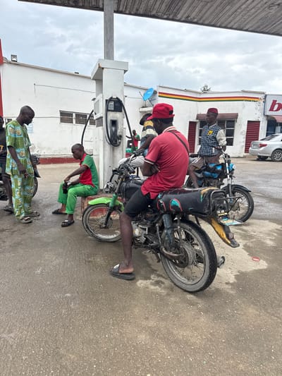 The image depicts a scene at a gas station in Lagos, Nigeria, under an overcast sky. In the foreground, a man is seated on a black motorcycle, facing away from the camera, wearing a red t-shirt and black shorts. The motorcycle has a yellow fender extension and a black rear cargo box. To the left, another man in a green and blue patterned traditional outfit stands near a motorcycle. Further left, a man in a green and red uniform is seated on a concrete block, holding a brown bottle. Behind him, another person in a similar uniform is visible. In the background, a white building with red and yellow trim stands behind a gas pump. A silver car is parked to the right. The ground is wet and appears to be paved. Several other motorcycles are parked around the gas station.