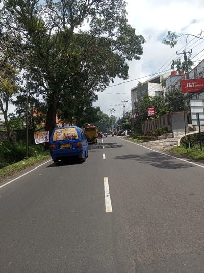The image captures a daylight street scene in the city of Ura-Dozhi, Bhutan. An asphalt road, marked with a broken white center line and a solid white shoulder line on the right, extends into the background under a partially cloudy sky. In the left lane, a blue minivan with license plate F 1213 MU is observed moving away from the viewer. The minivan's rear window displays a graphic containing multiple figures and partially legible text including "IYUNG GANI," and a banner with "ADALAH" is affixed to its side. A yellow dump truck is partially visible directly behind the blue minivan. Further ahead, multiple motorcycles are seen on the road. The left side of the road features a stone wall, dense green tropical foliage, and large trees, some hosting epiphytic plants. A white banner with red and yellow text, including the word "ADALAH," hangs from a pole near the road on the left. On the right side of the road, multi-story buildings are visible. One prominent building displays a red and white "J&T Express" sign, with another smaller red sign featuring white text positioned below it. Utility poles carrying numerous power lines run along the right side of the road, alongside a blank white signpost. No clearly discernible individuals are present in the foreground or midground.