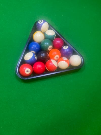 The image shows a rack of billiard balls on a green felt pool table. The balls are arranged in a triangular formation, with the cue ball at the front and the other balls filling the triangle. The balls are of various colors and numbers, typical of a standard pool set. The lighting suggests an indoor setting, possibly a game room or a bar. The scene is static, with no people or activity evident, focusing solely on the arrangement of the balls. There are numbers visible on some of the balls: 4, 13, 5, 9, 12, 10, and 3. The green felt of the table is textured, with some specks visible, possibly dust or lint. The rack is made of dark plastic and is positioned in the lower left portion of the frame. The background is a plain green surface, indicating the remainder of the pool table. No specific time of day or weather conditions can be inferred from the image. The context of Hof HaCarmel Regional Council, Israel, does not offer any visual cues within the image itself.