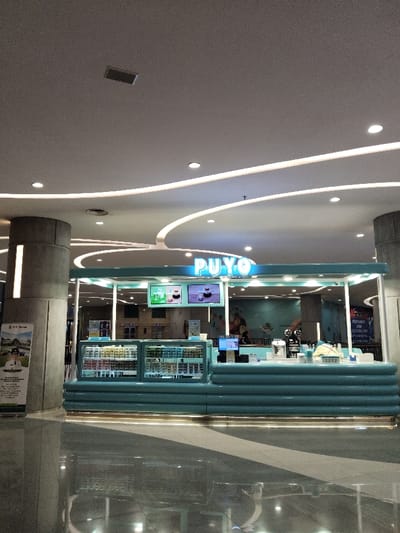 The image shows a food stall inside a shopping mall located in Telukjambe, Indonesia. The stall is decorated in a light blue and white color scheme and is labeled "PUYO". There are two display cases filled with various food items. One display case is directly behind the counter, and the other is behind the glass doors. The stall is located on a tiled floor in a well-lit mall environment with a curved ceiling.  There are no people in the stall. The stall is situated in a shopping mall in Telukjambe, Indonesia, at the coordinates -6.302 latitude and 107.278 longitude.  The stall is empty and appears to be ready to serve customers.