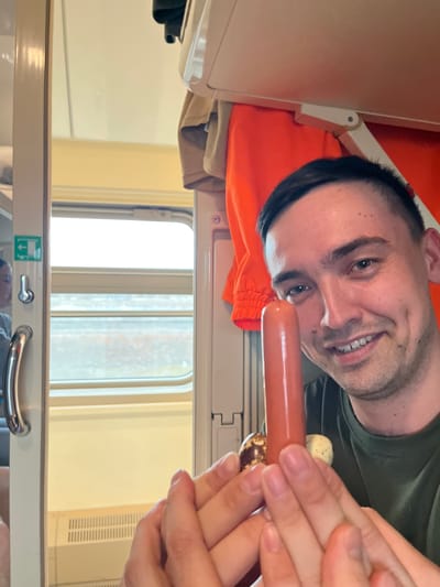 A man is pictured from an unusual angle, seemingly from the top bunk of what appears to be a Russian train, due to the style of window and bunk construction. He is smiling and holding a sausage, next to a small jar of condiments. The background shows a train window with vertical slats. The location is not visible in the image, so it's impossible to confirm it's in Yus'ki, Russia.
