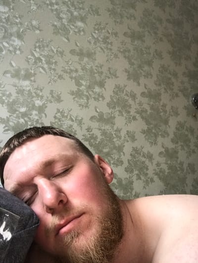 The image displays a close-up, overhead view of a light-skinned male sleeping or resting peacefully indoors in Sakmary, Russia. His eyes are closed, and his facial expression appears serene and relaxed. He has a prominent reddish-brown beard and mustache, along with short, dark hair visible on his head. His cheeks and nose exhibit a natural flush.

His head is gently resting on a dark grey or black fabric, which could be part of an item of clothing, a pillow, or a cap. A small, blurry, and illegible white mark, possibly text or a logo, is visible on this fabric near his chin.

The dominant background is a patterned surface, likely wallpaper or fabric, featuring a repeating floral design. This pattern consists of roses and leaves in muted green and grey tones, set against a lighter beige-grey base. The pattern covers the entire visible background, suggesting an indoor setting. The image primarily conveys a sense of calm and repose.