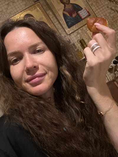 A woman is taking a selfie in a room with religious icons in the background. She has long, dark, wavy hair and is wearing a black shirt. She is holding a small piece of bread in her right hand and is wearing rings on her fingers and a bracelet on her wrist. The icons appear to be of Jesus and possibly other religious figures. The overall impression is of a personal moment captured in a culturally significant setting. The location is identified as the city of Turinsk, Russia.