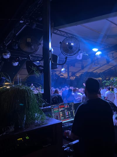 The image depicts an outdoor nighttime event at "The Forest" club in Beit Hanan, Israel. A DJ is visible from behind, operating a complex sound system with multiple screens and controls. The DJ is wearing a dark t-shirt and appears to be engaged in their work. In the foreground, to the left of the DJ, is a large potted plant with long, drooping green leaves. The background is filled with a crowd of people dancing and socializing under a canopy illuminated by blue and white lights. Several disco balls are hanging from the ceiling, reflecting the lights and adding to the festive atmosphere. Large fans are mounted on the structure above, likely for ventilation. The overall scene suggests a lively and energetic party environment.
