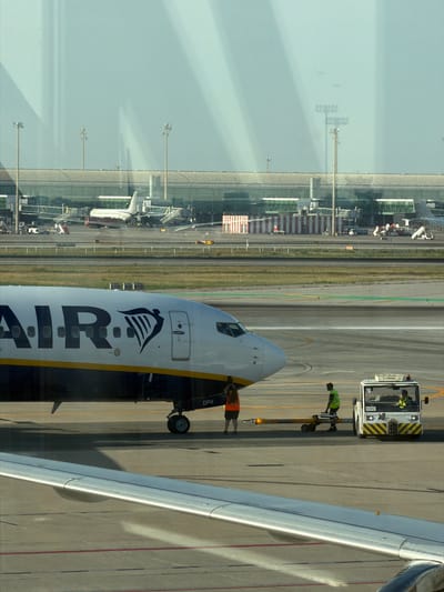 The image shows an airport scene at El Prat de Llobregat, Spain, as seen through a window. In the foreground, the wing of an airplane is visible, with a portion of the fuselage also showing. The fuselage has "RYANAIR" written in large black letters, along with the Ryanair logo. The airplane appears to be on the tarmac, with a tow bar connected to its front landing gear. A small white tug vehicle with yellow and black stripes is attached to the tow bar, with two people inside. Another person wearing an orange vest stands near the front of the airplane. In the background, the airport terminal building is visible, with jet bridges extending to other aircraft parked at gates. Several other airplanes are visible at various locations on the tarmac and in the distance. The sky is bright and overcast.