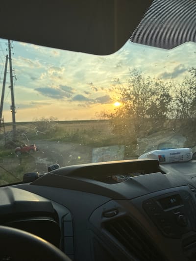 This image, taken from inside a vehicle in Mlekarevo, Bulgaria, captures a rural landscape at sunset. The view through the windshield shows a dusty road leading into a field under a sky filled with scattered clouds and the setting sun. On the left, a power line pole stands prominently. A small red object, possibly a toy car, is visible on the side of the road. The vehicle's dashboard is in the foreground, with a pack of cigarettes in a storage compartment and a pack of "Freshmaker" wet wipes on top. The sun's rays are casting a warm glow across the scene, creating lens flare and highlighting dust particles on the windshield. The overall impression is one of a quiet, rural setting during a beautiful sunset.