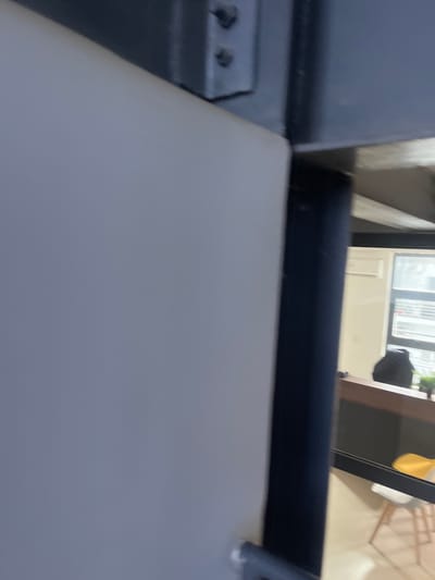 The image shows a close-up of a light gray surface in the foreground, possibly a wall or a door, with a dark blue, cylindrical object to its right. Behind this, a dark metal beam with visible bolts is present. Further in the background, through an opening, a room is visible with a dark desk or counter, a black backpack, a chair, and a window with blinds. The environment appears to be indoors, with artificial lighting. There are no people visible in the scene. The location of Karum, Nigeria, is not identifiable from the visual information provided in the image.
