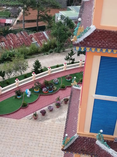 The image provides an aerial view of a temple complex in Lộc Quý, Vietnam. The foreground features the intricate, tiled roof of a temple, adorned with decorative elements in green and gold. The temple building itself is painted in light orange with large, blue-slatted windows visible. Adjacent to the temple is a paved courtyard with a repeating geometric pattern.
The middle ground showcases a well-maintained garden area with artificial green turf and red tiled pathways. Several potted plants with colorful flowers are arranged along these paths, adding vibrancy to the scene. A small, white statue of an animal is also present in the garden. A white, ornate railing encloses this garden area, separating it from a road or pathway beyond.
The background reveals a more rustic landscape, with sloping terrain covered in reddish-brown soil, likely agricultural fields. Several buildings with corrugated tin roofs, some appearing rusted, are scattered across this area. Tall green trees and dense foliage are interspersed throughout the landscape, suggesting a hilly or mountainous region. The overall impression is of a serene, spiritual site nestled within a semi-rural setting. No people are visible in the image.
