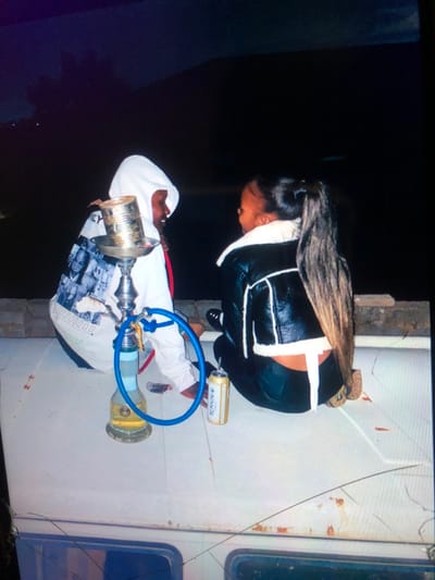 Two individuals are seated outdoors at night on a light-colored flat surface in Soweto, South Africa. The individual on the left, wearing a white hooded sweatshirt with black and white photographic prints and visible red undergarments, is barefoot and faces to the right. A silver-toned hookah, featuring a blue hose and a gold-colored repurposed metal can as its bowl, is placed on the surface in front of this individual. The individual on the right, wearing a black leather jacket with white shearling trim and black pants, is also barefoot and faces to the left. This person has long, dark hair styled in a high ponytail. Two aluminum beverage cans, one distinctly labeled "BERNINI" with an inserted straw, rest on the surface. A small dark object is visible near the hookah's base. The background is dark, with a faint, elevated landform discernible in the distance. The seating surface exhibits minor scuff marks and localized rust.