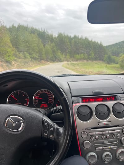 The image depicts the interior of a Mazda car while driving. The dashboard shows a mileage of 35056.1 km and a temperature of 36.6 degrees Celsius. The radio is tuned to FM2 100.00, and the time is 19:12. The driver is on a road surrounded by a forest, and the sky is overcast. The location is not in Dolno Drianovo, Bulgaria, and the information in the prompt is not contained in this image.