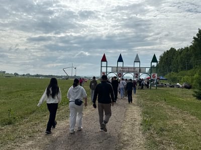 The image depicts a group of people walking along a dirt path in an open, grassy field. The sky is overcast with a dramatic cloud formation. In the background, there is a large, decorated archway with colorful triangular peaks and banners that appear to have text in Cyrillic. There are also some trees visible to the right of the archway, and more trees and fields in the distance. The scene suggests an outdoor event or gathering. The presence of the archway with Russian text indicates it is likely located in Russia. Based on the provided context, the location is identified as Старое Субханкулово, Russia.