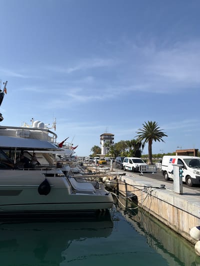 This media file depicts a scene at a marina, likely in Calvià, Spain, under a clear blue sky with wispy white clouds, suggesting a sunny day. The perspective is vertical, presenting a view from a yacht looking towards the shore and other vessels.
The primary subjects are several large yachts docked along a concrete pier. The yachts are modern and well-maintained, with some showing flags, including what appears to be a British flag and a red flag. Several white service vans are parked along the pier behind the yachts. A prominent palm tree stands tall among other lush greenery, adding a tropical touch to the setting. In the distance, a tower-like structure with white and brown accents is visible.
The activity appears to be a normal day at the marina, with people visible on one of the yachts, suggesting they are passengers or crew members preparing for or disembarking from a trip. The water is calm and reflects the surrounding structures and sky.
Notable details include the bright sunlight casting shadows, indicating it is likely daytime. The presence of multiple yachts and service vehicles suggests a busy port catering to luxury boating. The clear sky and calm waters contribute to a serene and pleasant atmosphere.
Visible text includes markings on the vans, such as "MTU" and what appears to be a French flag emblem on one of them. There are also some faint numbers visible on the pier itself, possibly dock numbers.