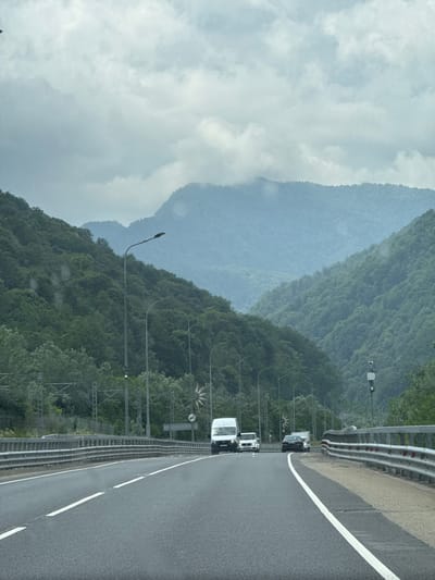 The image shows a view from a car on a highway in Chvizhepse, Russia, looking towards a mountain valley under a cloudy sky. The highway has multiple lanes separated by white lines and a metal guardrail on the right side. Several vehicles are visible on the road, including a white van in the left lane, a white car behind it, and a dark sedan in the right lane. The mountains on either side of the valley are covered in dense green trees, and the peaks are partially obscured by mist. Streetlights are spaced along the side of the highway. In the background, there are some power lines and structures along the left side of the road. The overall atmosphere is damp and overcast, suggesting recent or ongoing rain.