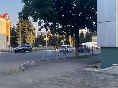 The image depicts a street scene in Antratsyt, Ukraine, likely during the daytime. A grey SUV is driving on a paved road, bordered by a decorative metal fence. Behind the fence, several cars, including a white hatchback and a dark sedan, are also on the road. A large tree with dark green leaves casts a shadow over the scene. To the left of the road, a building with a yellow facade and a red-tiled roof is visible. The sky is clear and blue. Several pedestrians are seen walking along the sidewalk behind the fence. Traffic signals and street signs are present, indicating a regulated urban environment. The overall impression is one of a typical street in a town or city.