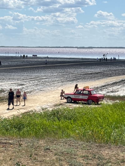The image shows a bright, sunny day at what appears to be a salt lake in Бурсоль, Russia. In the foreground, a patch of tall, green grass is visible. Behind the grass, a dirt path leads towards a pink-hued salt lake. Several people are walking on the beach and in the shallow water of the lake. A red and white car, with a "maxim" logo on its side, is parked on the dirt path. A woman is sitting on the trunk of the car. The sky is blue with scattered white clouds. The overall scene depicts a recreational area with people enjoying the natural environment.