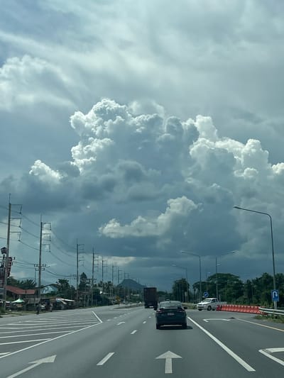 This image captures a highway scene in Ban Ko Pru, Thailand, under a dramatic sky filled with towering cumulus clouds. The perspective is from inside a vehicle, looking forward along a multi-lane asphalt road. In the foreground, white lane markings, including a large directional arrow and dashed lines, guide traffic. A dark sedan is visible in the right lane, followed by a white pickup truck in the adjacent lane. Further ahead, a semi-trailer truck is in the left lane.

The roadside features a row of utility poles with overhead power lines stretching into the distance. On the left side of the highway, a line of buildings, including small shops with awnings and residential structures, is visible. Green vegetation and palm trees are interspersed with these buildings. On the right side of the road, more utility poles and streetlights line the pavement, with a barrier of orange traffic cones and a blue sign indicating a U-turn ahead. The background shows a range of hills or mountains under the overcast sky. The overall atmosphere is somewhat somber due to the heavy cloud cover, with the bright white of the clouds creating a strong contrast against the darker storm clouds. No people are clearly discernible in the image.