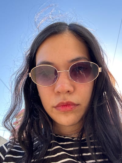 A woman with dark hair wears sunglasses. The sunglasses are round with gold frames. The sky is visible in the background. She is wearing a black and white striped shirt. Her lips are a natural pink color. The image is angled upwards, giving a slightly distorted perspective.