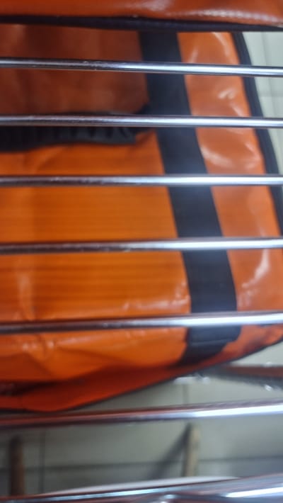 The image shows a close-up of an orange and black bag with metal bars in front of it. The bag appears to be made of a shiny, possibly vinyl or synthetic material, and has a pattern of horizontal orange sections separated by black stripes. The metal bars are thin and reflective, and are positioned horizontally across the frame, obscuring parts of the bag. The background is out of focus and appears to be a light-colored, possibly a wall or a partition. There are no people or specific identifiable landmarks visible in the image that would allow for determination of its location within the city of Megion, Russia. The image is a still shot and captures a static scene.