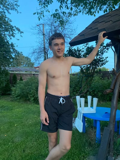 In a backyard in Uhornyky, Ukraine, a young Caucasian male stands outdoors near a wooden play structure. He is shirtless and wearing black athletic shorts with white trim. His right arm is extended and resting on the roof of the play structure. To his right, a bright blue plastic table with four white chairs is visible. The surrounding environment includes lush green grass, trees with dense foliage, and a wooden fence in the background. The sky is a clear, dusky blue, suggesting it is either early morning or late evening.