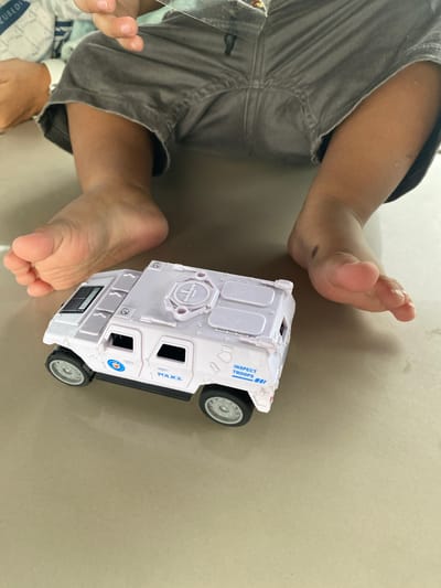 In Geulanggang Teungoh, Indonesia, a young child, partially visible, sits on a flat, light-colored surface, possibly a floor or table. The child wears gray shorts and is barefoot. Their legs and feet are in the foreground. A white toy police vehicle, detailed with "POLICE" and "INSPECT TROOPS" markings, is positioned in front of the child's feet. The scene is captured from a high-angle perspective.