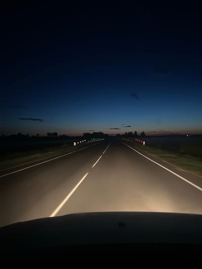 The image shows a view from a vehicle driving on a road at dusk or dawn. The sky is a deep blue, transitioning to lighter shades near the horizon, with a few faint clouds visible. The road ahead is illuminated by the vehicle's headlights, with white lane markings clearly visible. Red and white reflective markers line the right side of the road. The landscape on either side of the road is dark and appears to be rural, with silhouetted trees and fields. The foreground shows the dark silhouette of the vehicle's dashboard or hood. The scene captures a moment of travel in a rural Belarusian setting, likely on a highway, given the road markings and reflective markers.
