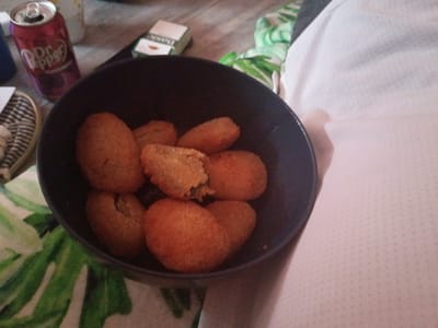 A slightly high-angle shot shows a dark-colored bowl filled with cooked, breaded food items resting on someone's lap. A can of Dr. Pepper and a pack of cigarettes are visible to the left of the bowl. Part of a striped item and a green and white patterned surface can also be seen in the lower left corner. The person's leg is covered by a white, textured fabric. The location, Clarksville, USA, is not deducible from the visual data provided.