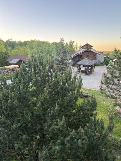 The image shows a view of a wooden building in Капцёўскі сельскі Савет, Belarus, partially obscured by trees. The building has multiple levels and a distinctive wooden design. The surrounding environment includes grass, other buildings, and a gravel pathway. The lighting suggests it might be near sunrise or sunset.