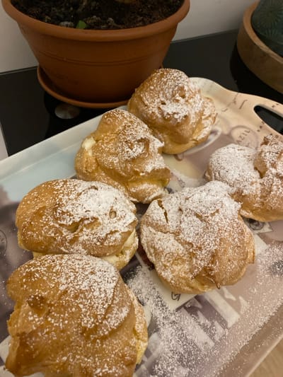 A tray holds several cream puffs, generously dusted with powdered sugar. The pastries are golden brown and irregularly shaped. A potted plant sits to the left of the tray, filled with dark soil. The tray features a patterned design and is resting on a black surface. The scene is brightly lit, highlighting the texture of the puffs and the sprinkling of sugar.