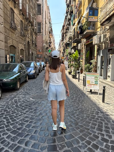 In a sun-drenched, narrow European street, paved with dark cobblestones, a young woman walks away from the camera. She is wearing a white tank top, light blue shorts, a white baseball cap, and sneakers. She is holding a smartphone in her right hand. The street is lined with tall buildings displaying various shops and businesses, with air conditioning units and exposed wires visible. There are parked cars, including a dark green vehicle in the foreground. A sign for "Farmact" and "Schettin" is visible on one building. Another sign in Italian advertises "PAGAMENTO UTENZE SCOMMESSE SPORTIVE" (Payment of Utilities Sports Betting) with logos for "mooney," "PUNTOLIS," and "SisalPay." The time of day appears to be midday, indicated by the strong shadows cast on the cobblestones. The scene suggests a casual stroll through a bustling urban environment in Naples, Italy, possibly on a warm, clear day.