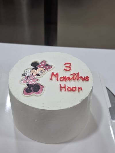 A close-up, high-angle shot shows a round, white frosted cake with a Minnie Mouse decoration and red lettering on top. The Minnie Mouse image is on the left side of the cake, showing her wearing a pink polka dot dress and bow. To the right of the image, red frosting spells out "3 Months Hoor". The cake is on a white surface, possibly a plate or cake board, with a portion of a silver knife visible on the right edge of the frame. The background is out of focus and appears to be a kitchen environment. The location of Dibba Province, Oman, is not depicted in the image.