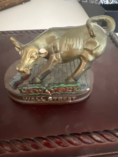 A bronze statue of a bull is displayed on a dark wooden surface. The bull is depicted in a charging stance, its head lowered and horns pointed forward. The base of the statue is circular and appears to be made of a different material, possibly painted to resemble cobblestones. The words "NEW YORK" and "WALL STREET" are inscribed on the base in raised lettering. The statue is golden-brown in color and has a glossy finish. The background is out of focus but suggests an indoor environment with a white wall and possibly other objects. The image is taken from a slightly elevated angle, looking down at the statue. There is no indication of the city of Hof HaCarmel Regional Council, Israel in the image.