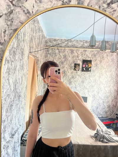 In this mirrored image, a person is taking a selfie. They are wearing a white tank top and black shorts. Their hair is braided and hangs over their shoulder. They are holding a pink phone in their right hand, using it to capture the reflection.

The background appears to be a room with textured wallpaper. On the wall, there are two small framed pictures of handprints. Hanging from the ceiling are three silver pendant lights. The lighting suggests it could be daytime, but the interior setting makes it difficult to determine. The overall mood is casual and personal. The image contains no visible text. The location context provided is Blizhnii Khutor, Moldova.