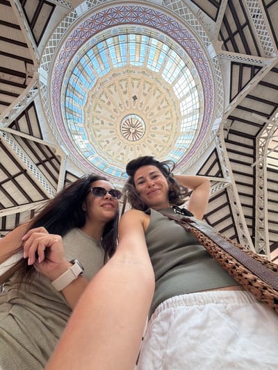 Two women are captured in a low-angle, wide shot selfie within a grand, domed interior space. The architectural setting features a large, circular stained-glass ceiling with intricate geometric patterns and decorative floral motifs, illuminated by natural light. The dome is supported by a complex network of white metal beams and trusses, creating a sense of spaciousness and grandeur.
The woman on the left, with long dark hair, is wearing sunglasses and a light olive-colored top. She is resting her arm on the shoulder of the other woman. A watch with a light-colored band is visible on her left wrist. The woman on the right, with dark brown hair, is smiling and has her right hand behind her head, resting on her arm. She is wearing a dark olive-colored ribbed tank top and white linen shorts. A woven leather and straw handbag is slung across her right shoulder. The overall mood appears relaxed and joyful.
The architecture suggests a public building, possibly a market or a covered public space. Given the context of Alboraya, Spain, this could be the Central Market of Valencia, which is known for its stunning modernist architecture, including a prominent dome and stained-glass windows.