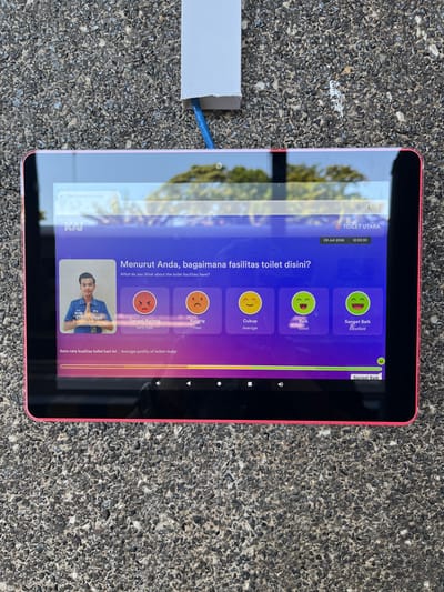 An outdoor eye-level medium shot shows a tablet computer mounted on a textured, gray, pebbled surface, likely a wall or sidewalk, in Desa Tambakreja, Indonesia. The tablet, with a bright pink casing, displays a digital survey about toilet facilities. The screen shows a photograph of a young man in a blue uniform, identified as Khairul Anjal, along with a question in Indonesian and English: "Menurut Anda, bagaimana fasilitas toilet disini? What do you think about the toilet facilities here?". Below the question are five emoji-based rating options: "Sangat Kurang" (Very Poor), "Kurang" (Poor), "Cukup" (Average), "Baik" (Good), and "Sangat Baik" (Excellent). A progress bar at the bottom of the screen indicates "Rata-rata kualitas toilet hari ini | Average quality of toilets today," with the "Sangat Baik" option highlighted. Above the survey, a date and time are visible: "09 Juli 2026 12:02:30" and "TOILET UTARA." A white electrical box with a blue cable exiting is attached to the pebbled surface above and to the left of the tablet. The background is blurred, showing green foliage and a bright sky, suggesting an outdoor, daytime setting.
