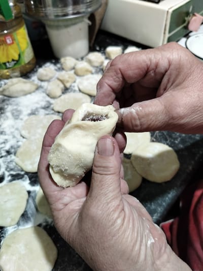 In this close-up shot, a pair of hands is in the process of making dumplings, likely vareniki, a traditional Ukrainian dish. The hands, showing signs of age with visible wrinkles, are carefully shaping a piece of dough that has been filled with a dark red substance, possibly berries or fruit jam. A dusting of flour coats the dough and the surface beneath, indicating a kitchen setting where dough is being prepared. Scattered around are several other round, flattened pieces of dough, ready to be filled and formed.

In the background, partially visible elements suggest a home kitchen environment. To the left, a bottle of cooking oil with a sunflower label is present, and beyond it, a stainless steel pot or container and a white canister. Further to the right, a part of a light-colored appliance, possibly a mixer or another kitchen gadget, is visible. The overall scene is dimly lit, suggesting it might be indoors during the day or evening. The focus is squarely on the hands and the delicate task of dumpling making, conveying a sense of domestic activity and tradition. There is no explicit indication of the time of day or weather conditions. The location context is Shchastia, Ukraine, which aligns with the cultural context of making dumplings. The emotion conveyed is one of focused, patient craftsmanship. No readable text is clearly visible in the image, aside from the label on the oil bottle which is largely illegible.