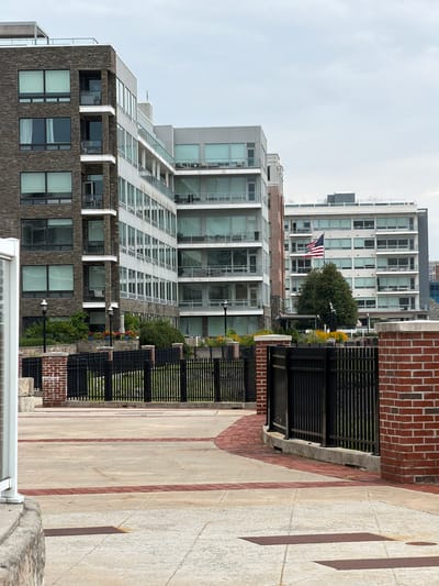 This image depicts a view of modern apartment buildings, likely in an urban or waterfront setting. The buildings are tall and feature a contemporary architectural style with numerous large windows. One building in the background has an American flag displayed, suggesting a location in the United States. The scene appears to be outdoors, with a paved walkway in the foreground. The walkway is bordered by a brick wall and a black metal fence. There are some potted plants and greenery visible along the fence line, indicating some landscaping. The sky is overcast, suggesting it might be a cloudy day. The time of day is difficult to determine precisely due to the lighting, but it appears to be daytime. There are no people visible in the image. The overall impression is one of a well-maintained urban environment. The location context provided is Edgewater, United States of America.