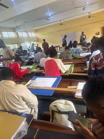 This media file shows a classroom setting in Amansee, Nigeria. The room appears to be an educational institution, possibly a university or technical college, with tiered seating arrangement typical of lecture halls. Several students are visible, engaged in what seems to be a class or workshop.
The subjects in the image are primarily people and classroom objects. The people are predominantly young adults, dressed in various casual and formal attires, suggesting a diverse student body. They are seated at desks, some are looking at whiteboards with diagrams, while others are focused on their work or personal devices. Objects include desks, chairs, whiteboards, pens, rulers, bags, and mobile phones. There are also ceiling fans installed on the ceiling, and windows with louvers allowing natural light to enter the room.
The activity appears to be a learning session, possibly a lecture or a practical class involving drafting or drawing, given the presence of drawing tools and diagrams on the whiteboards and desks. Some students are actively working on their projects, while others are observing. The presence of a mobile phone being used by one of the individuals suggests it might be a modern educational environment.
Notable details include the natural lighting, implying it is daytime. The setting is indoors, within a classroom. The overall mood seems to be focused and studious. There is some visible text on a wooden board that reads "Jerkibe." Additionally, a mobile phone screen displays some content, but it is not clearly legible. The room's architecture, with its windows and ceiling fans, suggests a tropical climate.